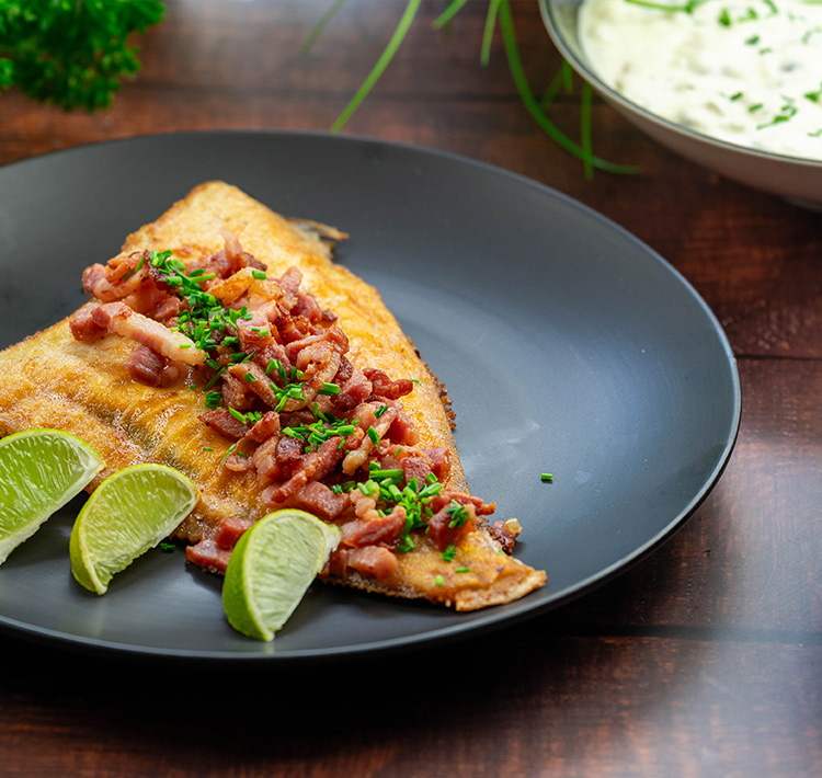 Angerichtetes Fisch-Essen auf schwarzem Teller mit Limettenscheiben und Speck.