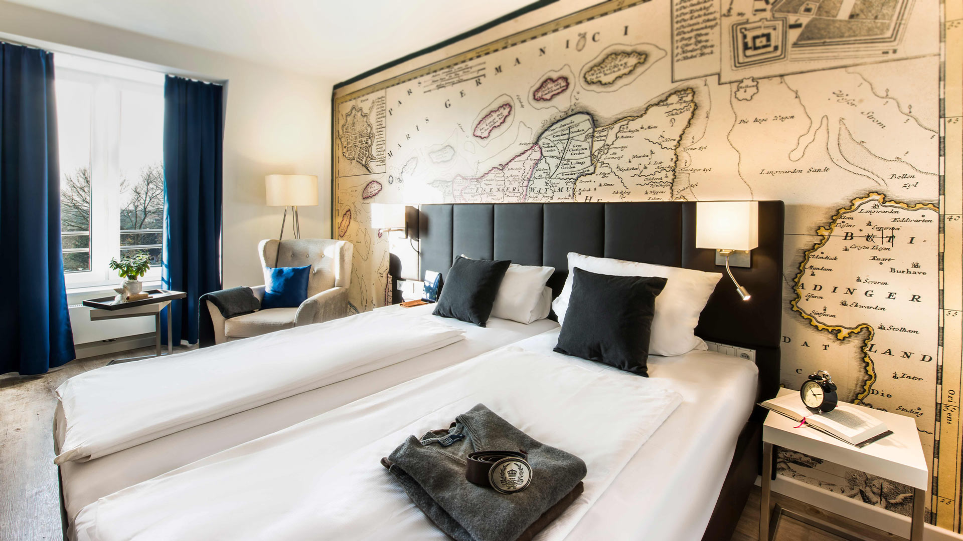 Nahaufnahme des Hotelbettes und einem Sessel im Hintergrund und einer Landkarte an der Wand.