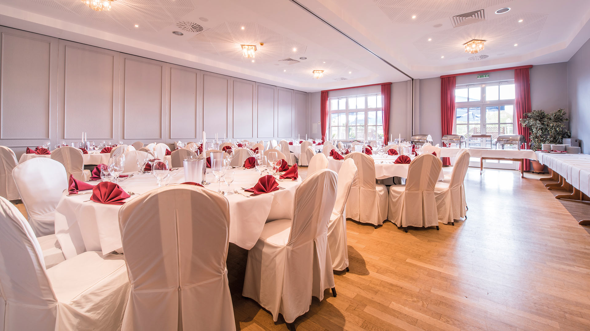 Raum mit eingedeckten, runden Tischen mit weißen Tischdecken und einem Buffet im Hintergrund.