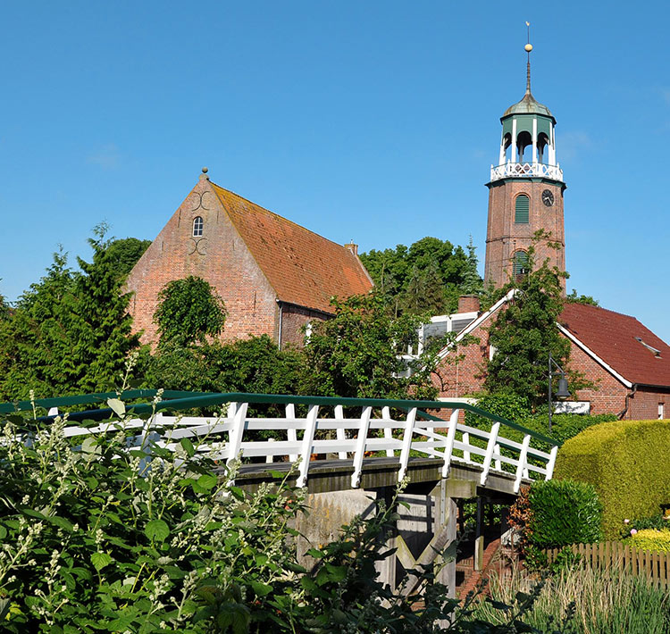 Eine Brücke verläuft zwischen Büschen zu Backsteinhäüsern.
