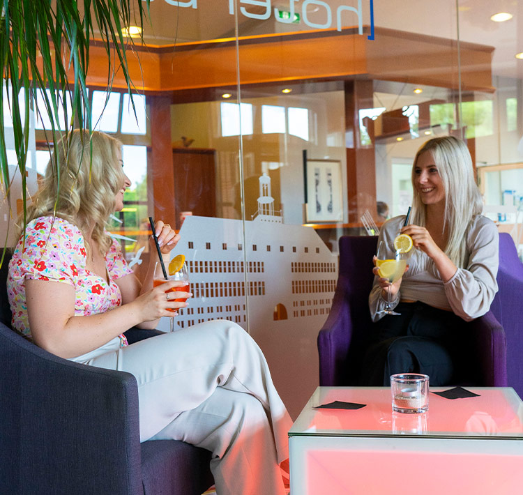 Zwei blonde Frauen sitzen mit Getränken in einer Lounge.