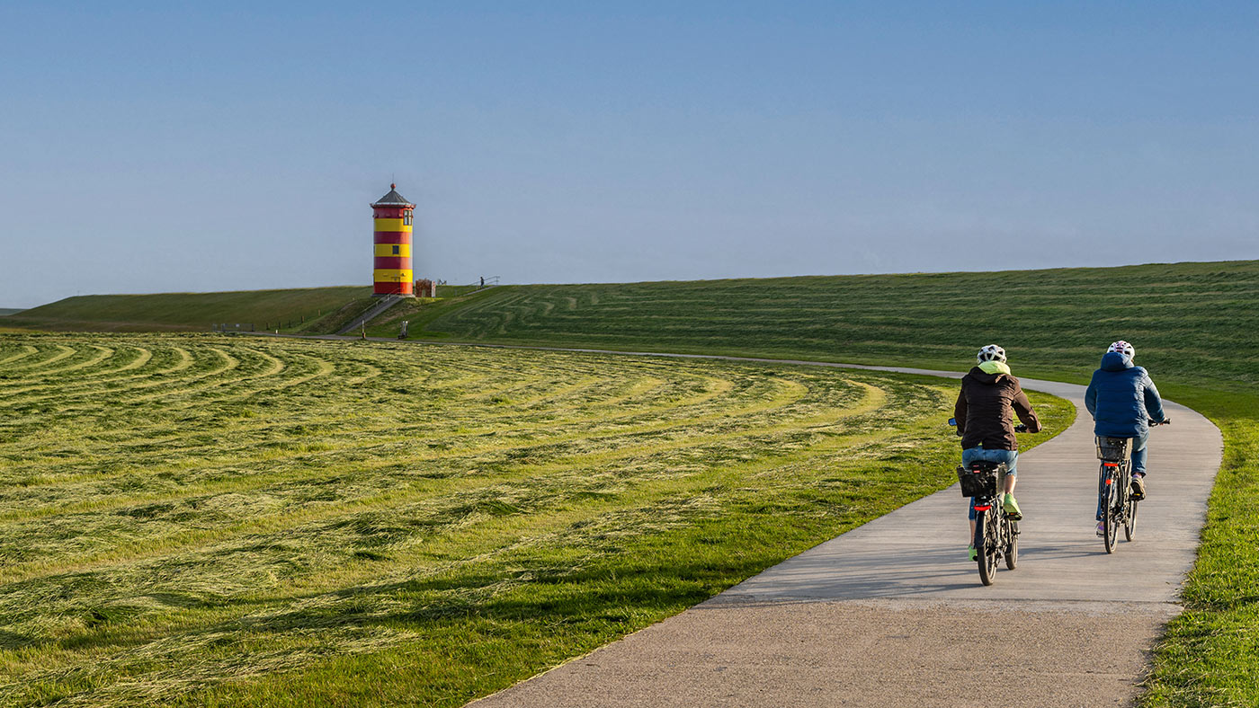 Zwei Fahrradfahrende auf einem Wiesenweg vor einem Leuchtturm.