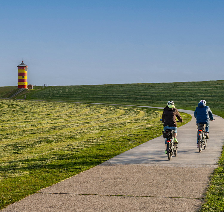 Zwei Fahrradfahrende auf einem Wiesenweg vor einem Leuchtturm.